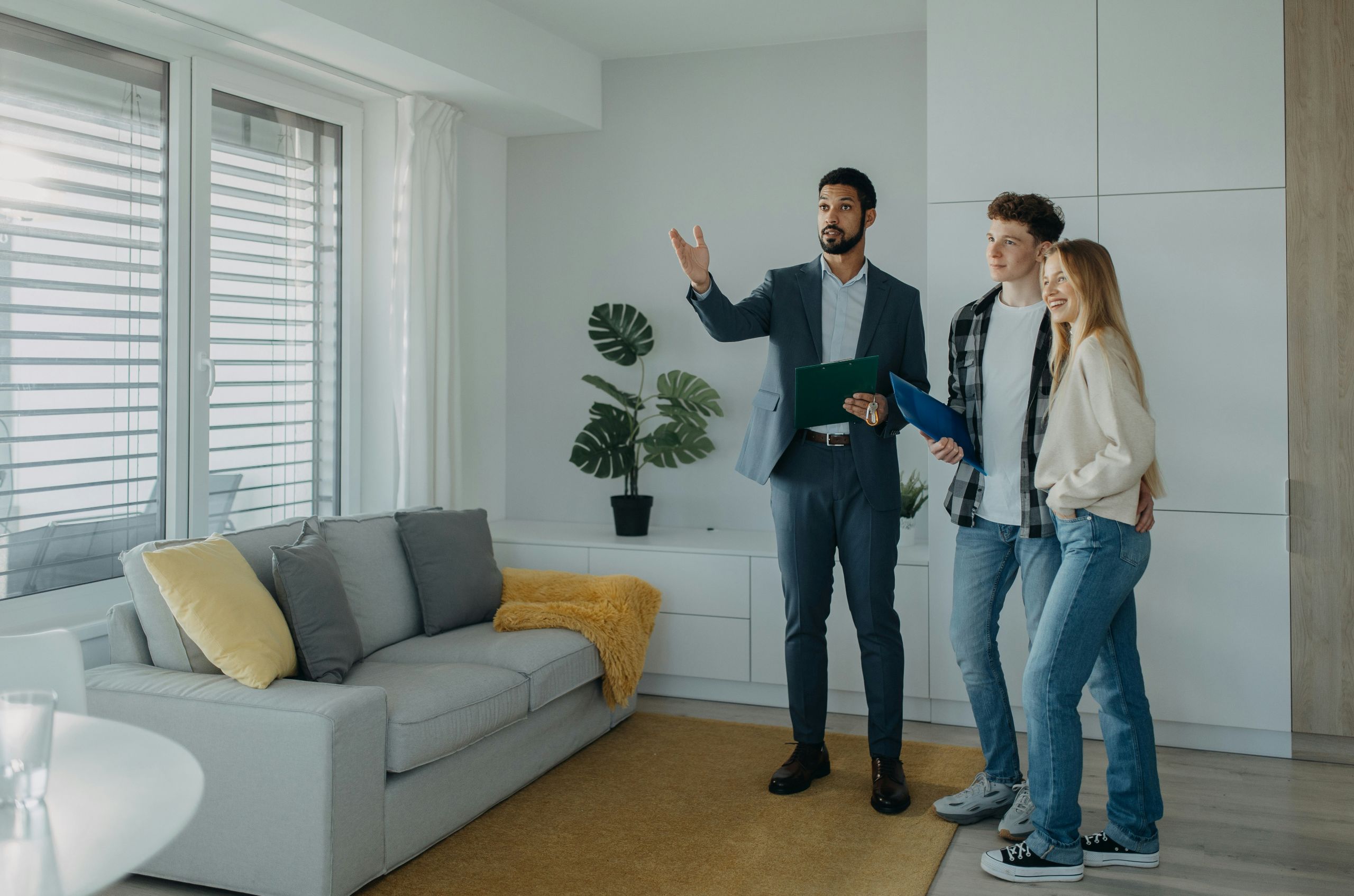 A real estate agent in a suit shows a modern living room to a young couple. The buyers stand together, smiling, while the agent gestures toward the bright space featuring a couch, plants, and large windows.