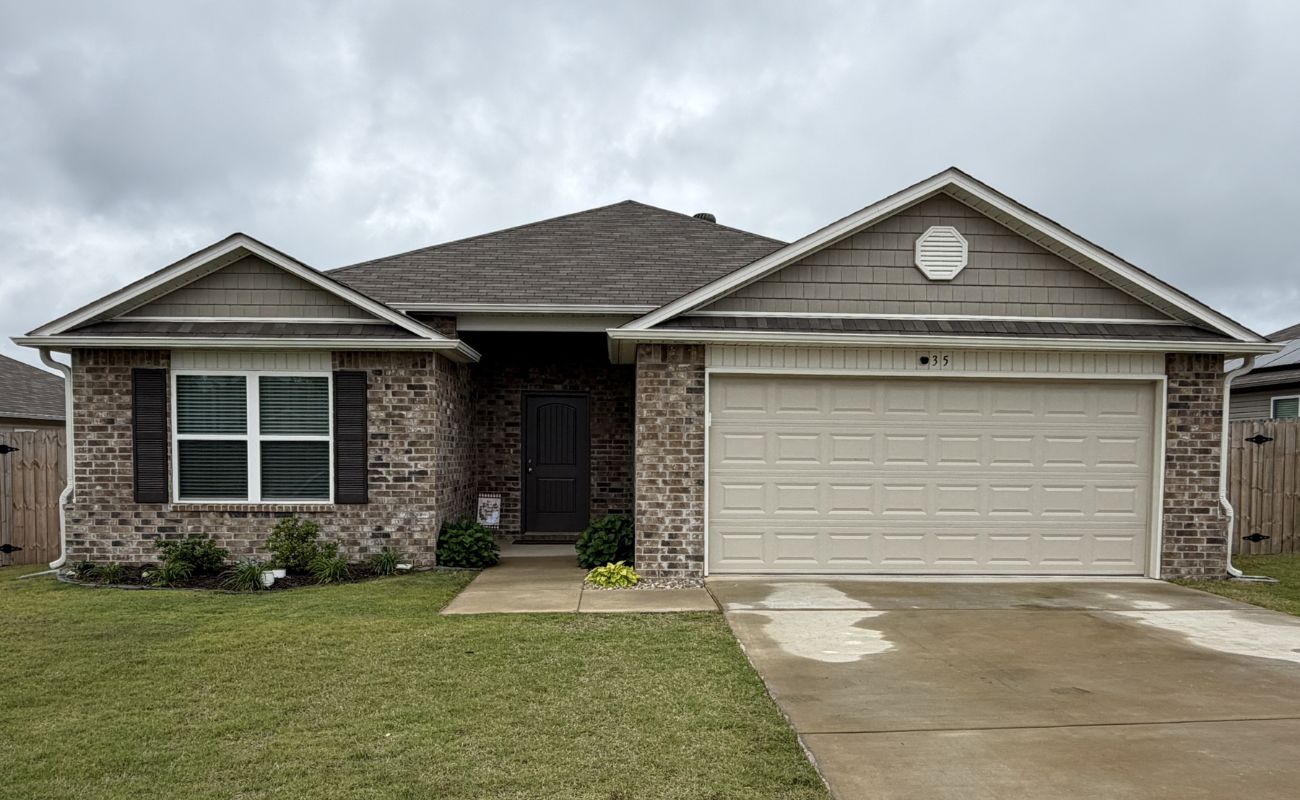 Single-story brick house with a two-car garage, brown shingle roof, black front door, and fenced backyard. The lawn is neatly trimmed and the sky is overcast.