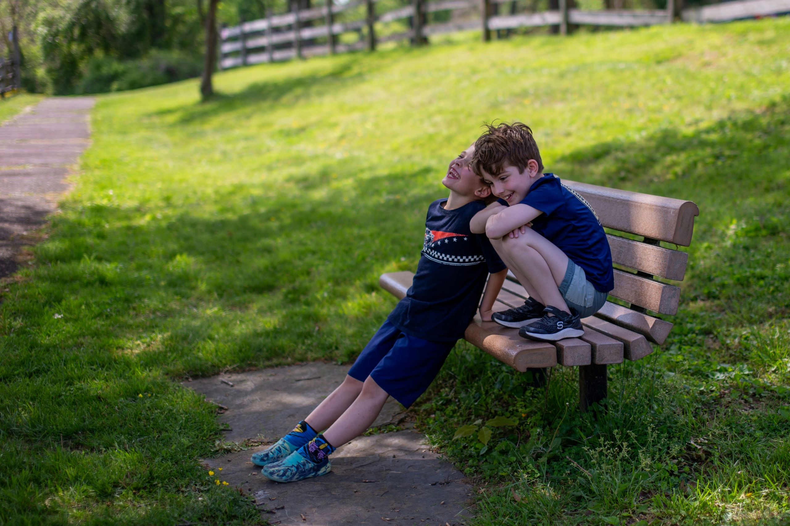 Two young boys sit on a park bench in the sun, talking excitedly about moving to Arkansas. One leans back, smiling, while the other sits with arms on his knees; both are dressed in navy shirts and shorts, surrounded by green grass and trees.