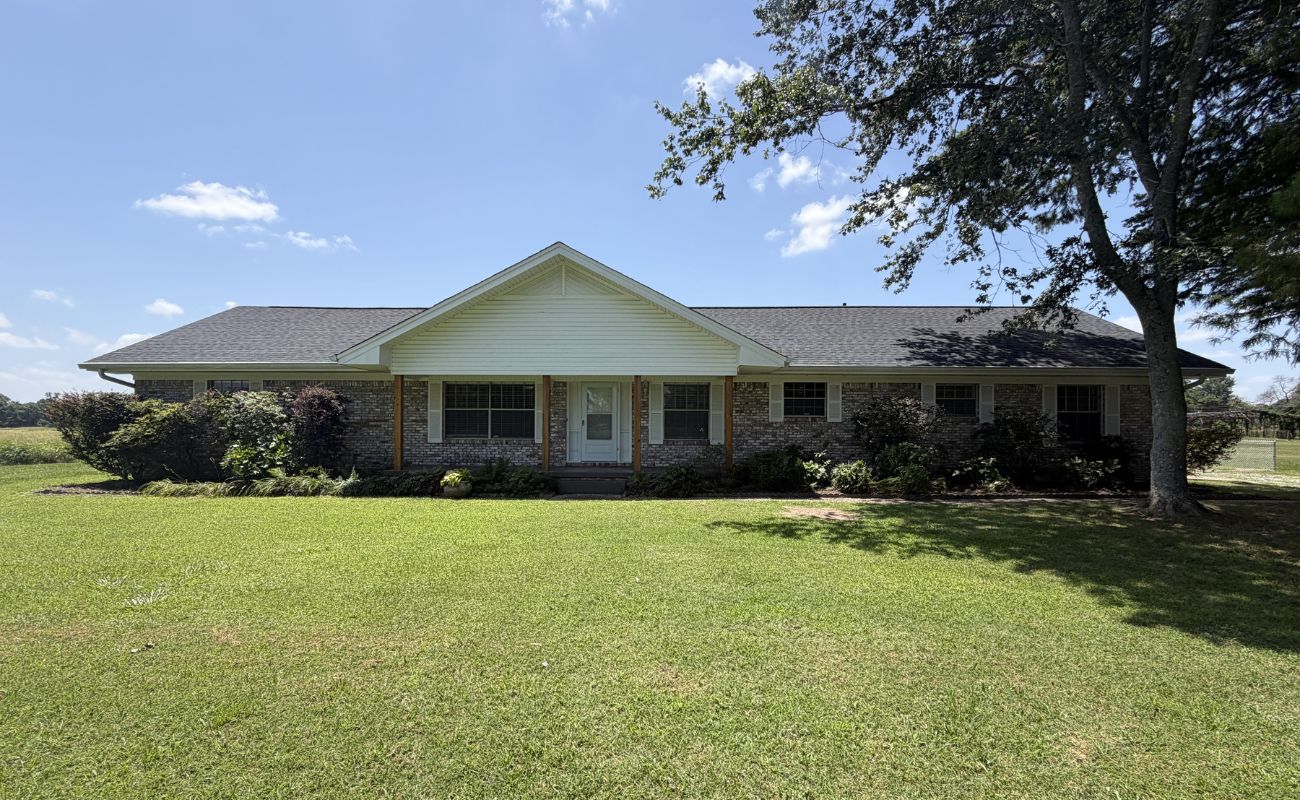 Single-story brick house with white trim, a centered front door, and a large, grassy lawn. Bushes and trees surround the front, and the sky above is mostly clear and sunny.