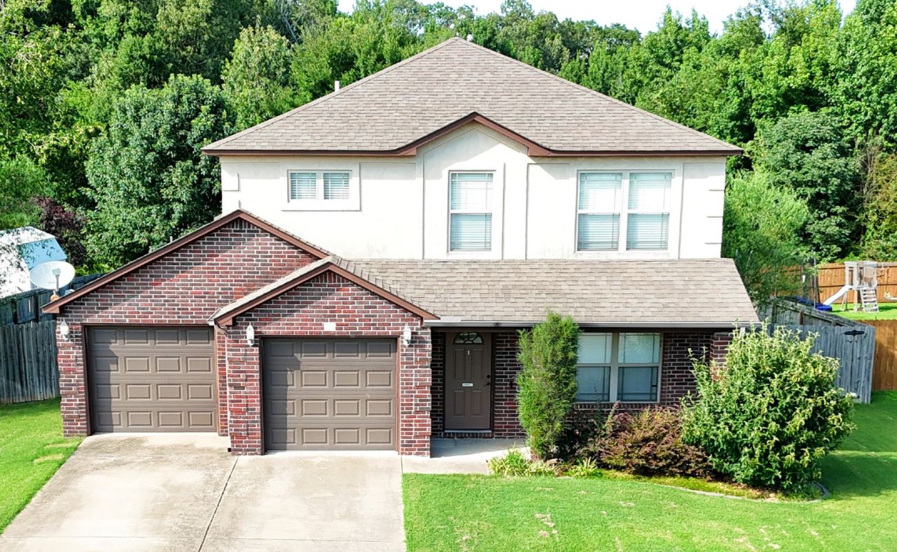 A two-story suburban house with a brick and stucco exterior, two-car garage, brown doors, front lawn, and surrounded by trees and a wooden fence.