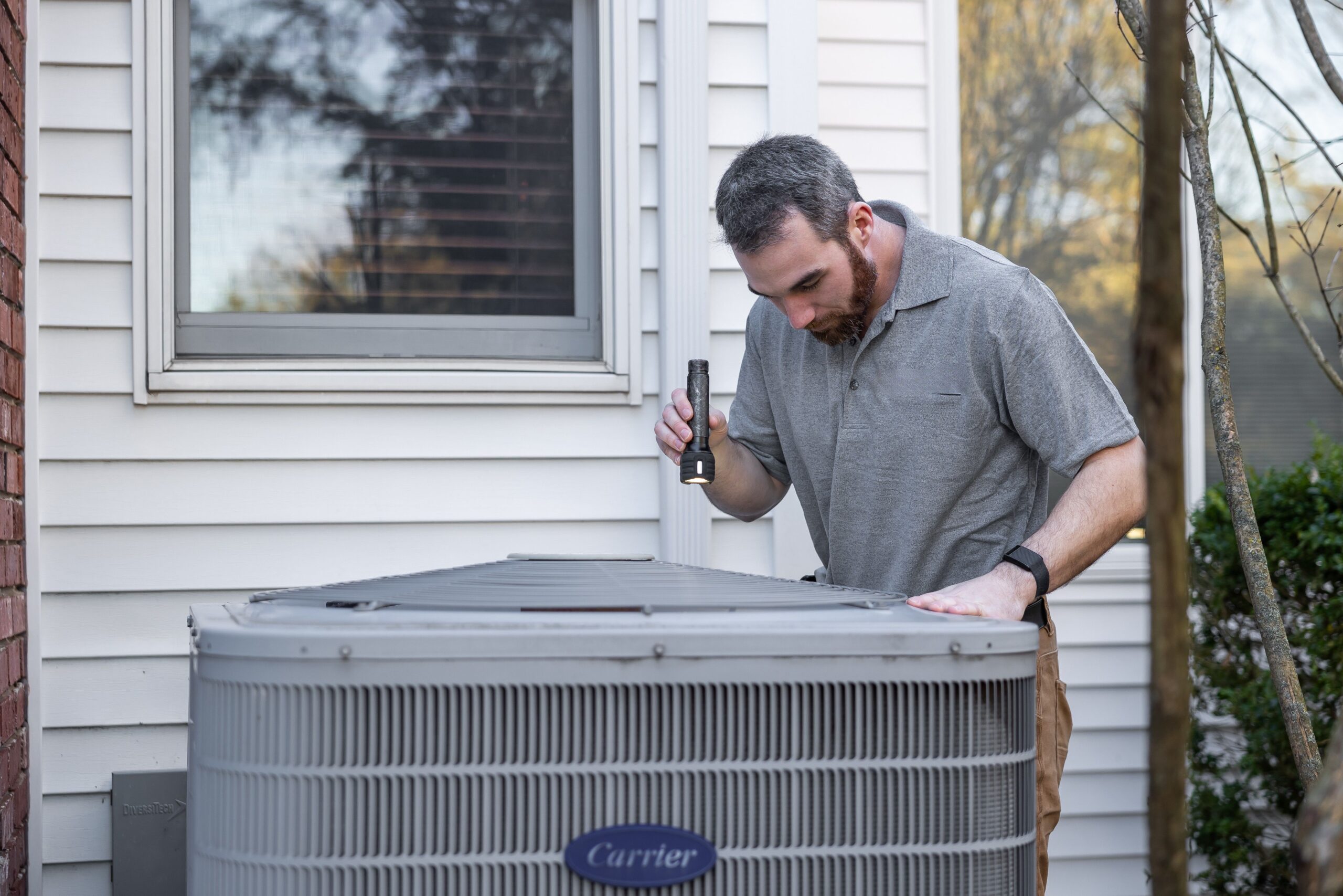 A man inspects an outdoor air conditioning unit with a flashlight near a house window during the daytime, an essential step on any seasonal home checklist in Central Arkansas to help prevent foundation problems caused by Arkansas clay soil.