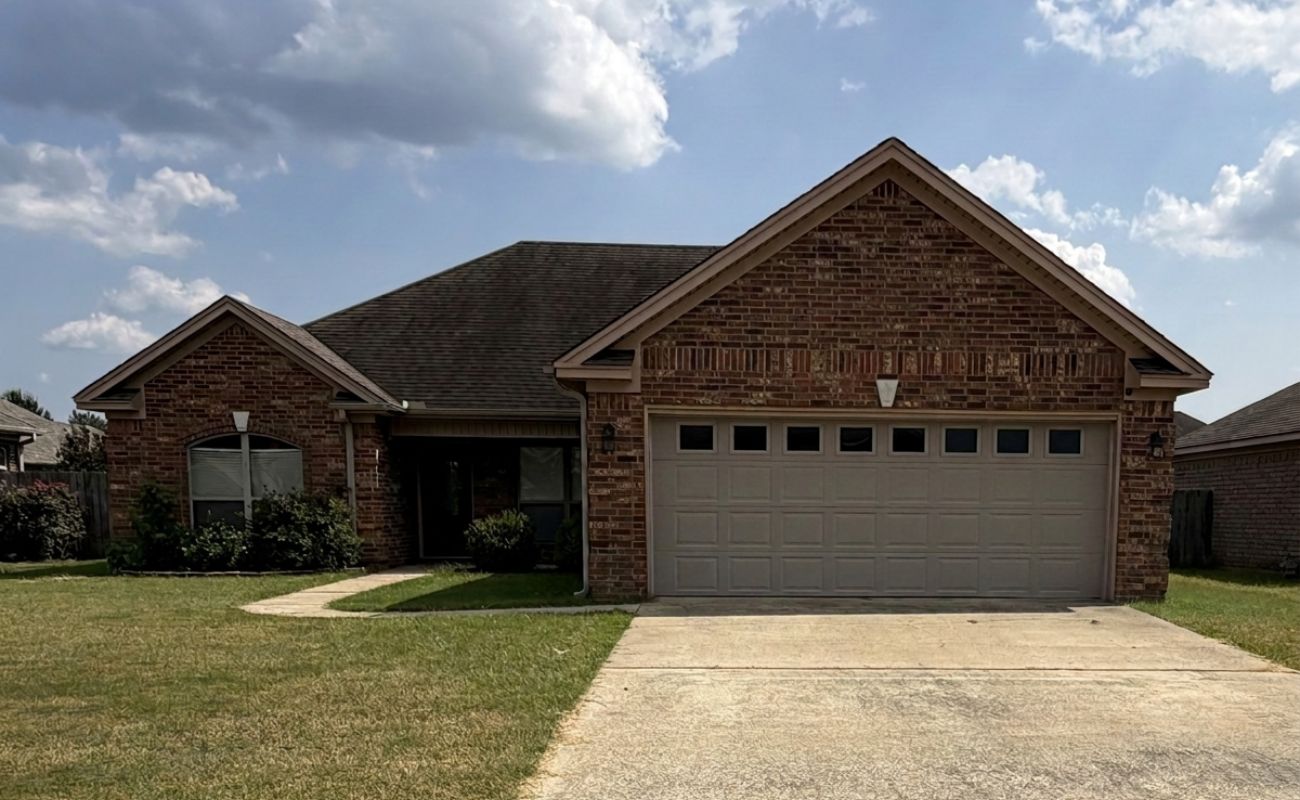 A single-story brick house with a two-car garage, a front lawn, concrete driveway, and partly cloudy sky overhead.