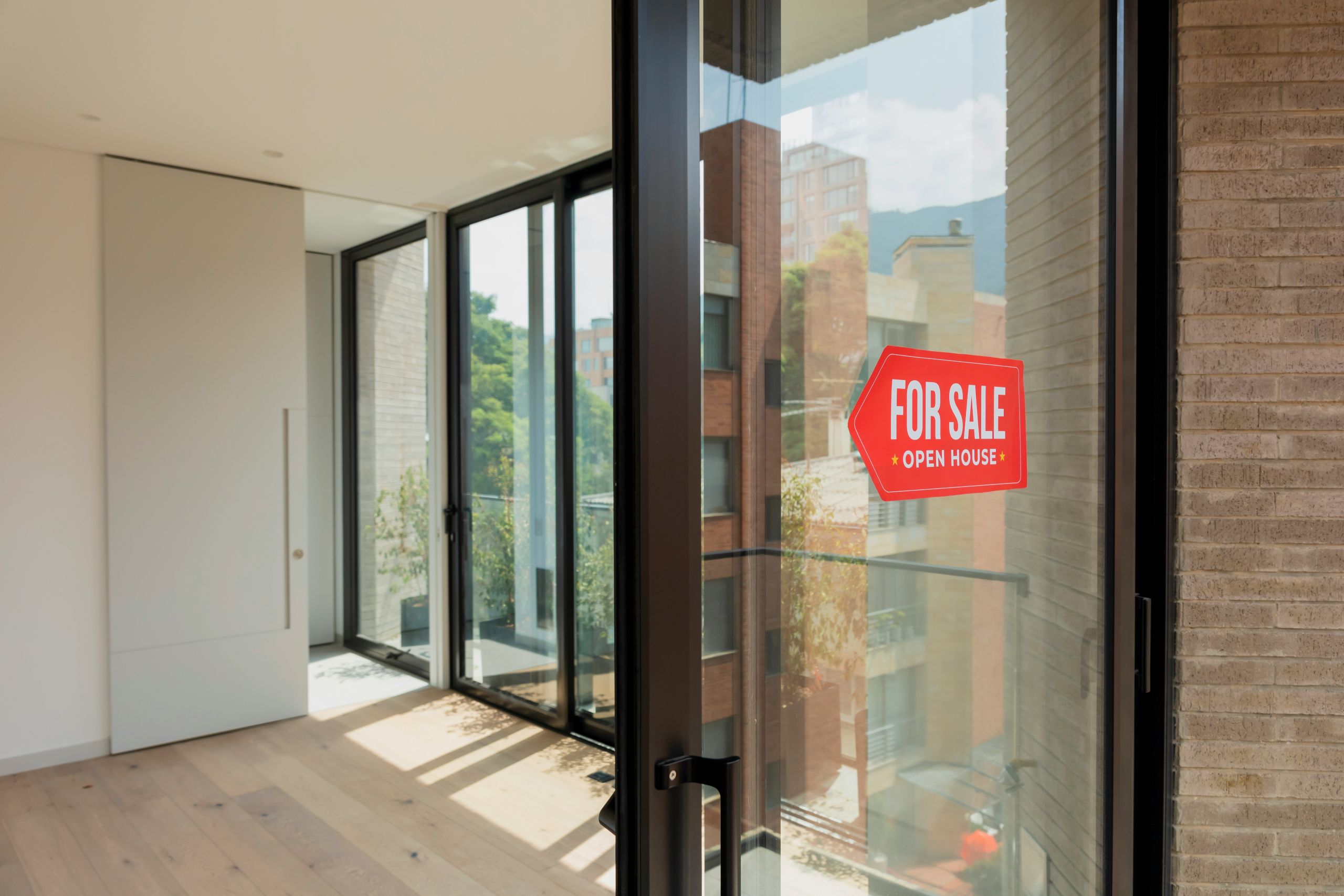 Bright, modern apartment interior with large windows and a glass door displaying a red For Sale Open House sign. City buildings and green trees are visible outside.