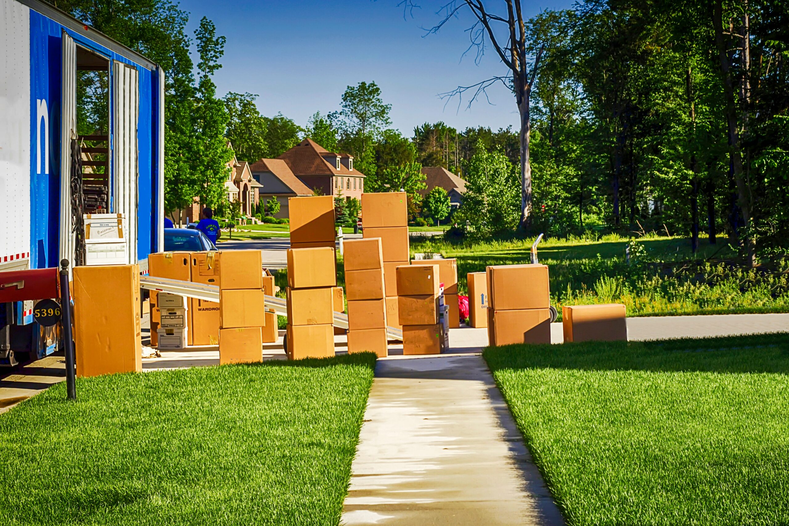 A sidewalk leads to a street where numerous cardboard boxes are stacked near a moving truck, with green grass and houses visible in the background—capturing a typical scene of moving day while living in Arkansas.