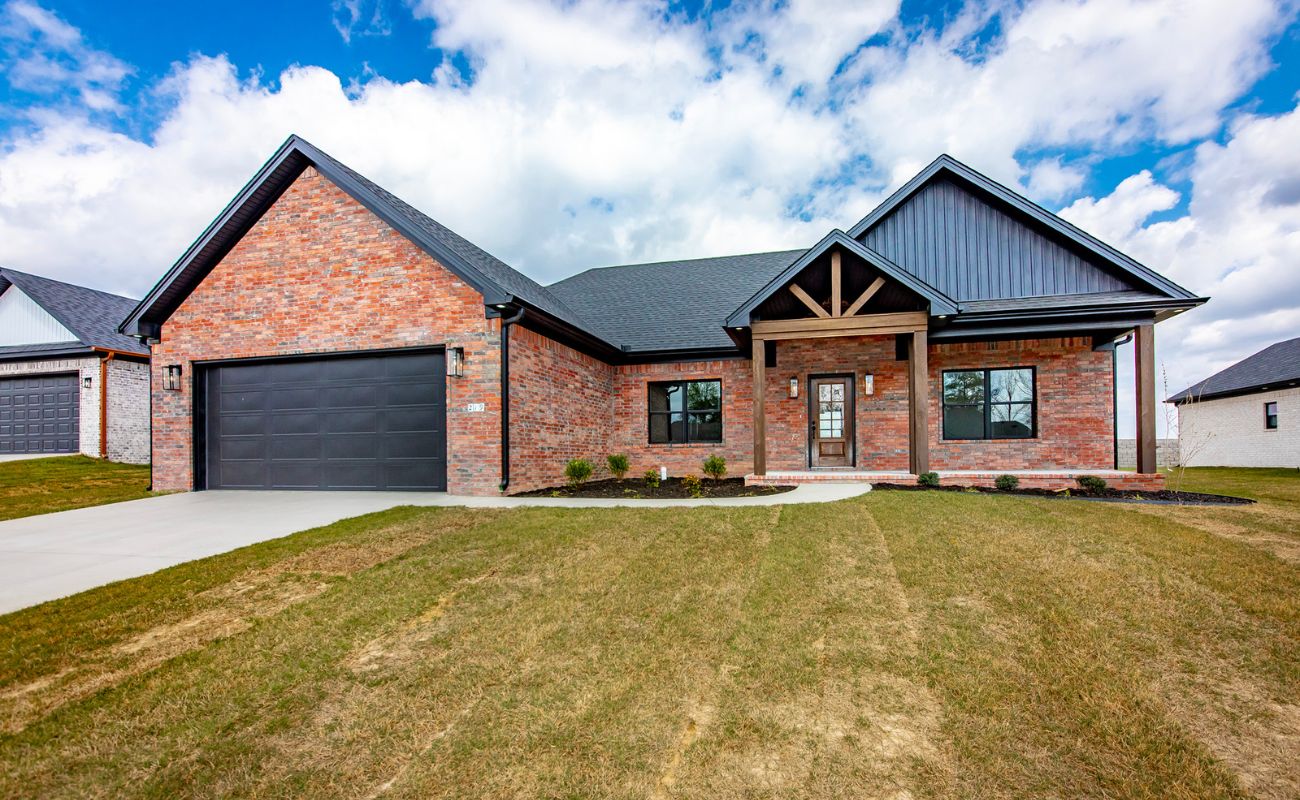 A modern brick house with a dark roof, large driveway, double garage, covered porch, and landscaped front yard under a partly cloudy sky.