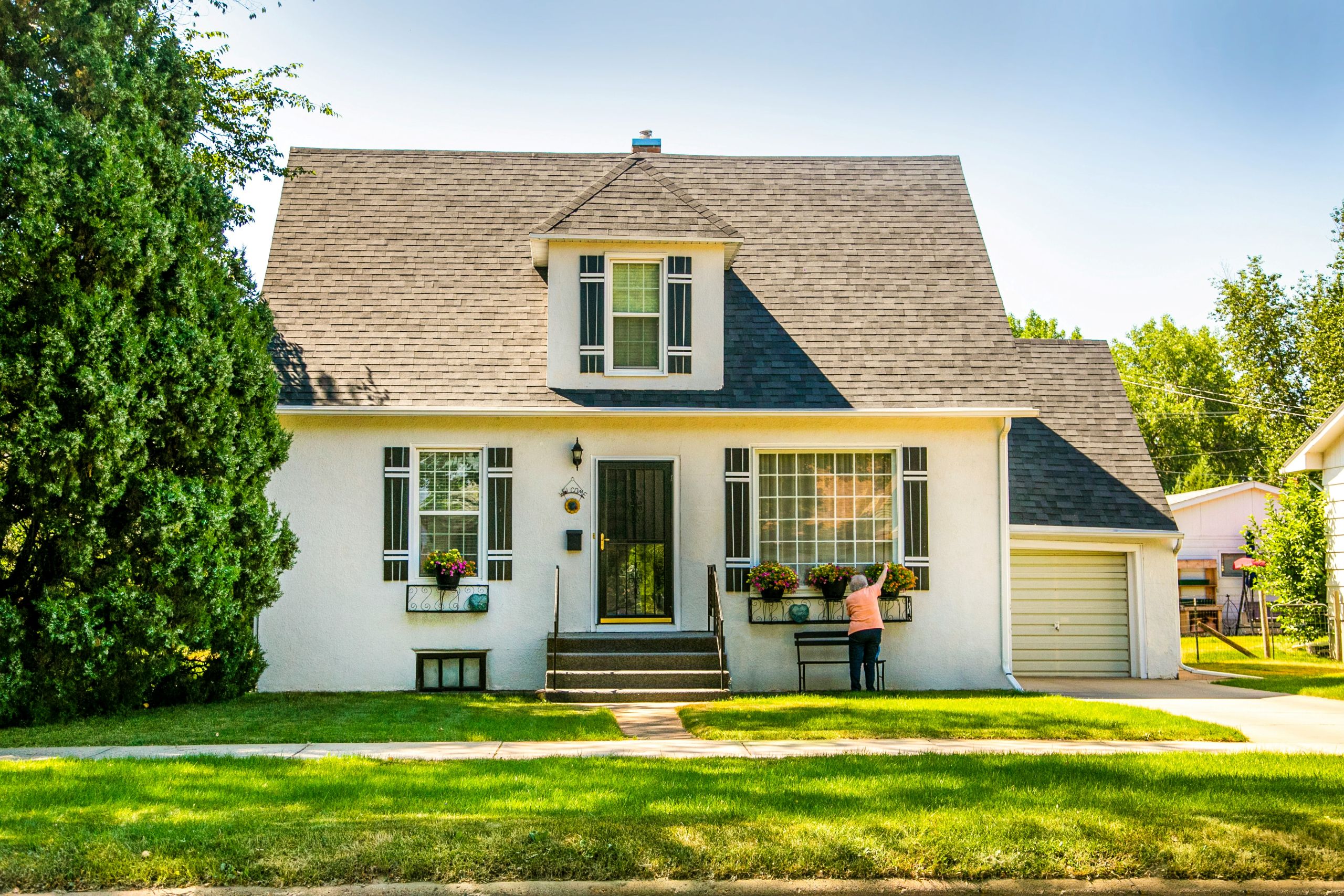 A person tends to flowers outside a white house with a steep roof and attached garage on a sunny day. The yard is green and well-kept, with large windows and shrubs.