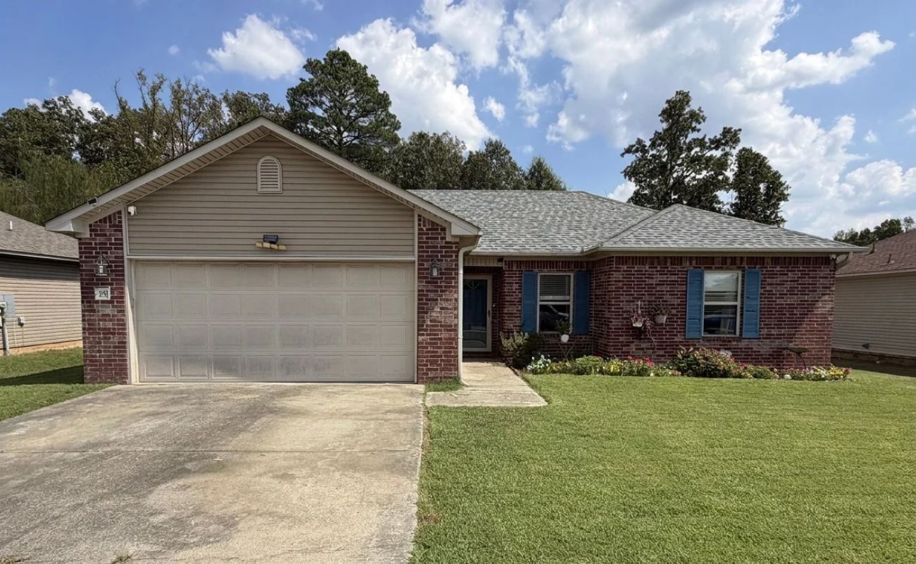 15 Castleton Drive Ward, AR A single-story brick house with a two-car garage, beige siding on the gable, and blue shutters on the windows.