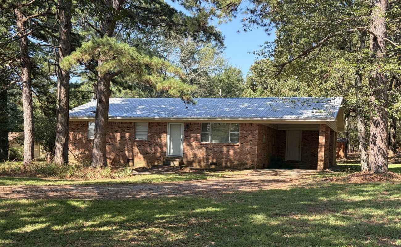 27217 Arkansas Hwy 107 Cabot, AR A single-story brick house with a metal roof and a carport, surrounded by trees.